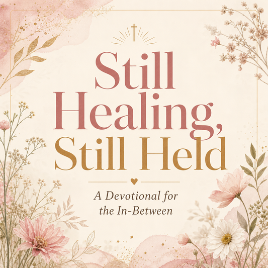 A square book or digital cover design with the title 'Still Healing, Still Held' in large serif text, plus the subtitle 'A Devotional for the In-Between.' The cover has a soft cream background with floral illustrations in blush pink, white, and muted green, plus small gold accents and a small cross above the main title.